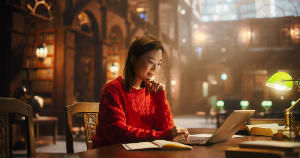 istock Thoughtful Multiethnic Asian College Student Using Laptop Computer to Study in a Classic Library. Beautiful Young Woman Learning Online, Getting Ready for Exams, Taking Notes and Drafting an Essay. 2160742885
