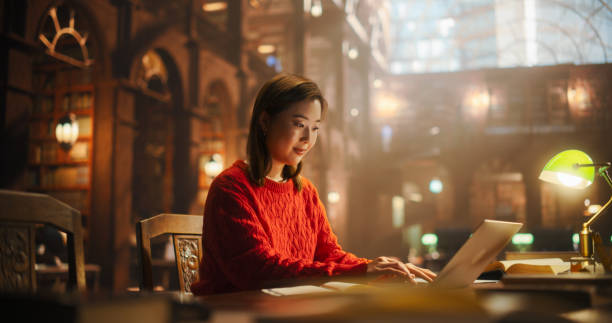 istock Focused Japanese Female Student Studying in a Classic Library, Using Laptop for Online Education, Surrounded by Books in a Historical Setting, Preparing for University Exams. 2160742905