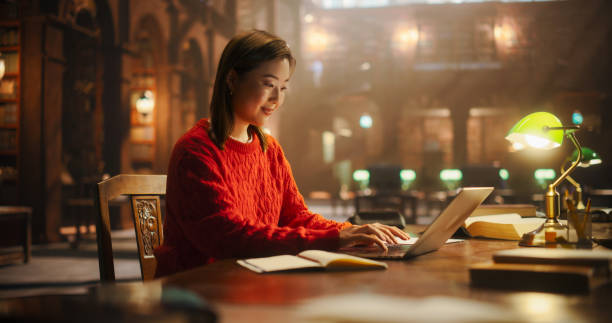 istock Focused Asian Female Student Studying in a Classic Library. Young Woman Using Laptop for Online Education, Surrounded by Books in a Historical Reading Room. 2160742913