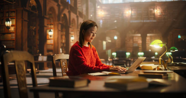 Dedicated Asian Female Student Engaged in Online Learning at a Classic Library, Using Laptop to Study and Prepare for University Exams Amidst Books and Historical Ambiance.