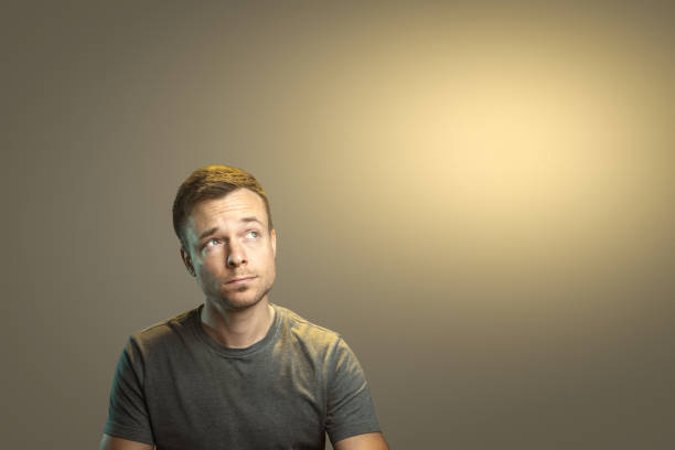 Upper body of a man in his early 30s in casual clothing is looking thoughtfully up to the side. He is isolated against a neutral background with ample copyspace.