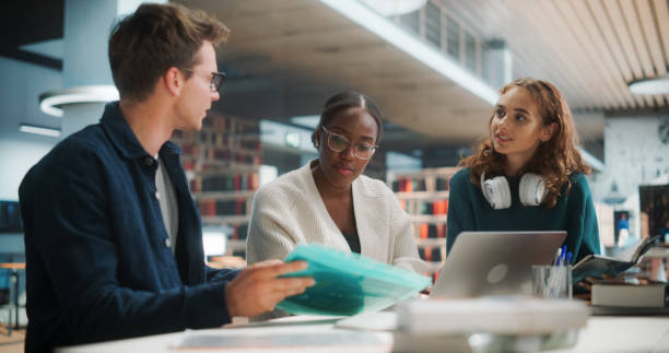 istock Diverse Group of Students Collaborating on a Project in a Modern Library. Young Black Female and Caucasian Peers Discussing Academic Materials and Using a Laptop for Research. 2160751387