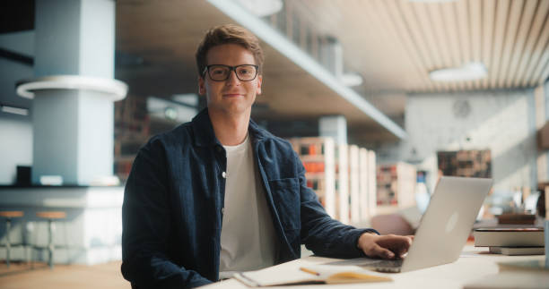 istock Confident Young Caucasian Male Student Working On a Laptop in a Modern Library, Surrounded by Books. Smart, Casual Outfit, Glasses, Engaged in Academic Research with a Focused Expression. 2160751490
