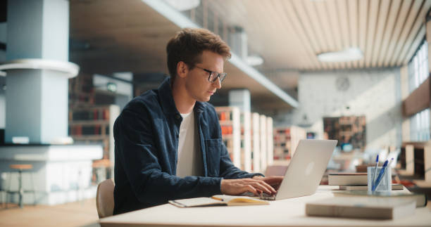 istock Young Man Studying in Modern Library. Caucasian Male Using Laptop Computer for College Research, Surrounded by Books and Academic Materials, Focused on Learning and Education in University Setting 2160751507