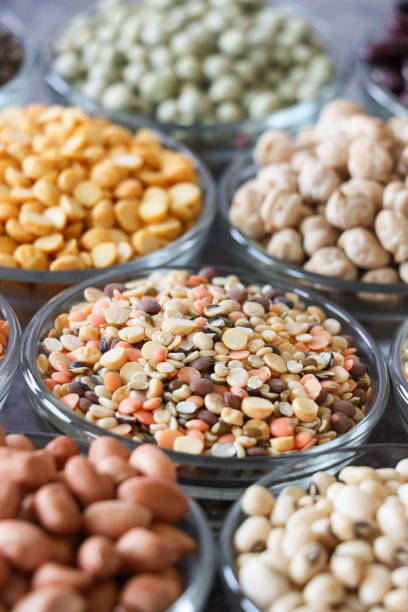 Stock photo showing close-up view of glass bowls containing piles of mixed split lentils, including red, yellow, brown and green lentils, red skin peanuts (Arachis hypogaea), also known as groundnuts or monkey nuts, dried black eyed peas (Rongi dal / white lobia), also known as black eyed beans, split yellow lentils, also known as Arhar dal or Toor dal, dried chickpeas (Cicer arietinum), also known as ceci beans, chana, chichi beans, garbanzo beans, gram or Egyptian peas, and dried green peas (vatana), also known as marrowfat peas or matar. All these seeds and pulses are considered to be very healthy foods, containing a rich source of carbohydrates, dietary fibre, protein, vitamins and minerals. grupos de imágenes en primer plano cuencos de vidrio llenos de montones de legumbres, nueces y semillas, lentejas partidas mixtas, cacahuetes de piel roja, guisantes de ojo negro secos (rongi dal / lobia blanca), lentejas amarillas partidas (gandules), ga