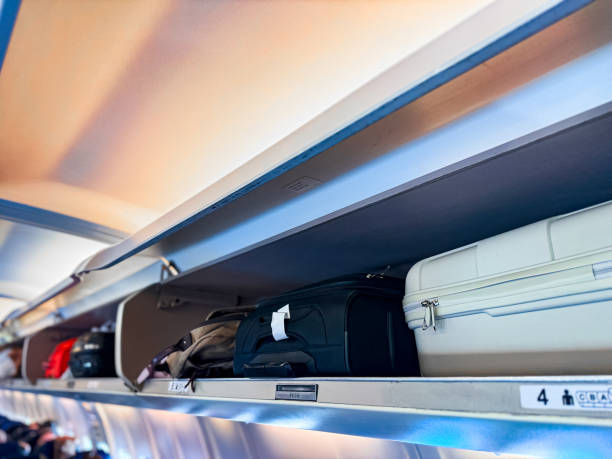 istock Overhead storage compartment inside airplane cabin filled with various carry on luggage, including black and white suitcases, backpacks, bags, with compartment doors open, visible seat row numbers. 2160762924