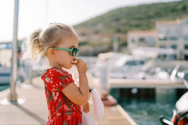 istock Little girl eats a bagel while standing with a toy rabbit on the pier. Side view 2160768317