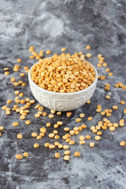 Image of dried yellow split pea, chickpea, chana dal lentils in white ceramic bowl, surrounded by scattered beans, peas and pulses, healthy food, nuts, seeds, against granite kitchen worktop background