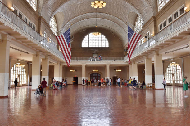 New York, USA - May 16 2023: A large room in Ellis Island with a flag hanging from the ceiling. People are standing and sitting in the room