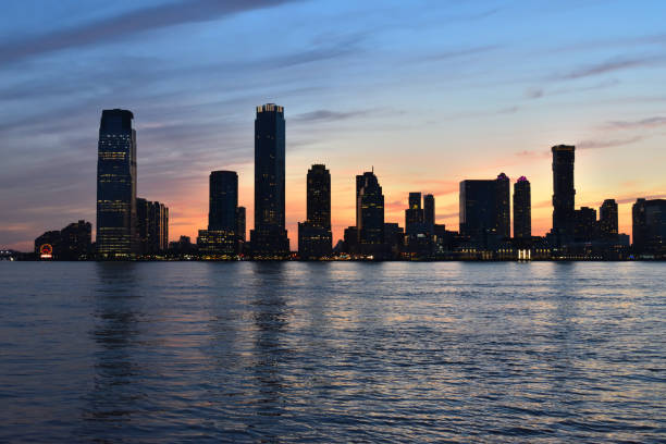 A city skyline is reflected in the water at sunset. The sky is a mix of orange and blue hues, creating a serene and peaceful atmosphere