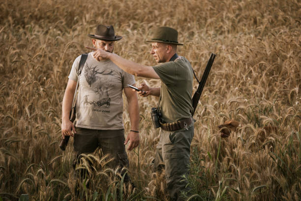 Two hunters stand in a field of golden wheat, looking at a mobile phone screen for navigation. The man on the right, wearing a green shirt and hat, points to the phone screen with his left hand, while holding a rifle in his right hand. The man on the left, wearing a beige t-shirt with a deer image and a brown hat, listens intently. The golden light of the setting sun bathes the scene, creating a warm and inviting atmosphere.