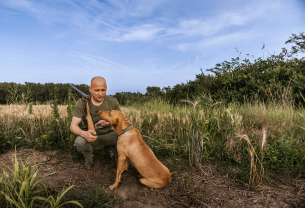 A hunter with a shotgun and his loyal dog stand together in a field, likely after a hunt. The sky is blue with a few clouds, and the grass is green.
