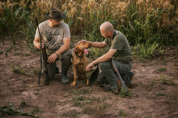 Two hunters, one with a hat, rest in a field after hunting. They have their rifles and a dog with them. The dog is sitting between the hunters and is being petted by one of them. The hunters are in a relaxed pose, but they are still prepared for the next hunt.