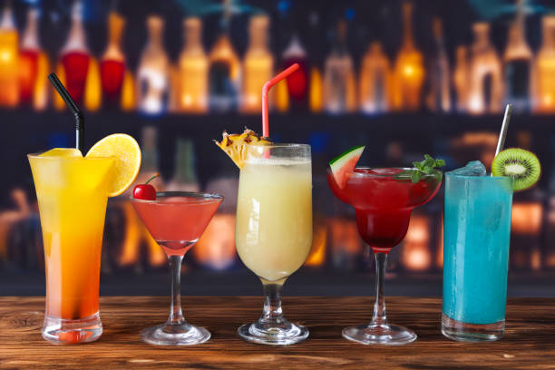 Image of row of alcoholic cocktail drinks with garnishes, tequila sunrise, cosmopolitan, piña colada, watermelon margarita, blue lagoon, drinking straw, bar counter, pub setting background, focus on foreground stock photo
