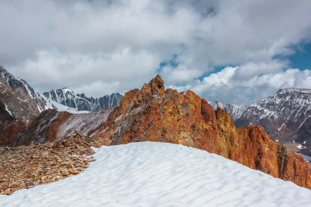 Snowfield on stony mountain ridge and vivid sharp pinnacle in sunlight. Shiny pointy peak of gold color. Colorful red rocky peaked top. Freshly fallen snow in high mountains. Sunny cloudy alpine view.
