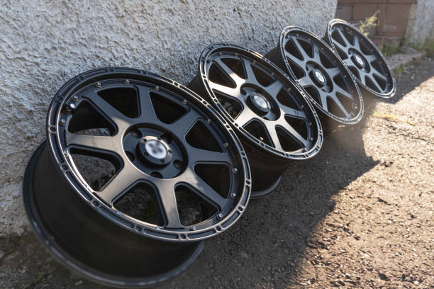 A set of glossy black alloy wheels leaning against a wall on a sunny day, casting shadows on the ground.