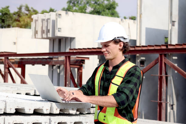 istock Side view of happy worker engineer with safety vest and helmet typing on laptop computer while inspecting working area at construction building site. Young architect works at building site workplace. 2160848573