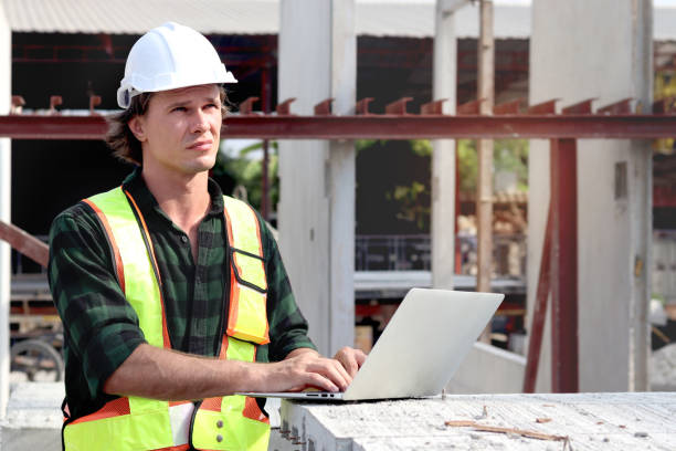istock Portrait of happy worker engineer with safety vest and helmet typing on laptop computer while inspecting working area at construction building site. Young architect works at building site workplace. 2160848699