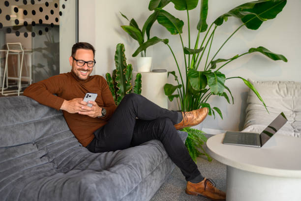 istock Mid adult professional texting over smart phone while relaxing on sofa by laptop in modern office 2160857178