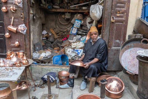 Fes, Morocco - March 23, 2024: An artisan skillfully shapes copperware in his workshop at Place Seffarine, a historic square known for its tradition of metal craftsmanship dating back to the 8th century.
