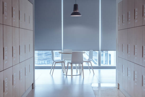 A modern office locker area with a sleek and minimalist design. The corridor is flanked by rows of wooden lockers, each labeled with white numbers. At the end of the hallway, there is a small seating area featuring a round table and white chairs
