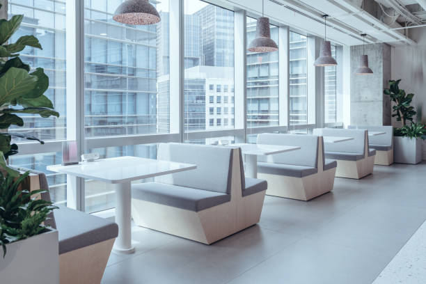 A modern and minimalist dining area with sleek, grey booth seating and white tabletops. The space is illuminated by natural light streaming through large windows, offering a view of adjacent office buildings.