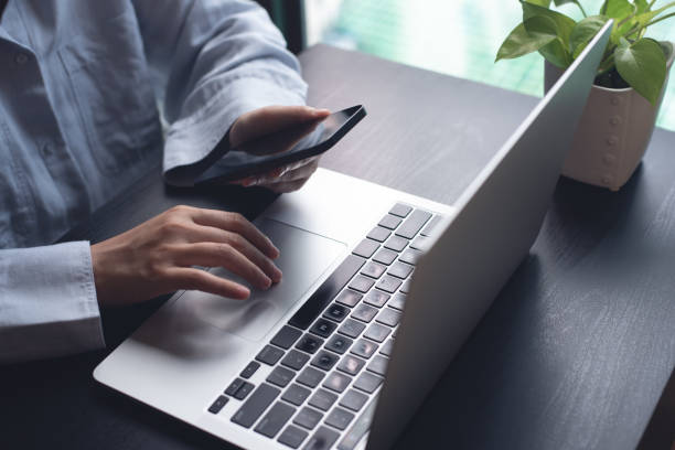 istock Business woman sitting at office table using mobile phone while online working on laptop computer at home office, business casual, telecommuting, working from home, close up 2160869152