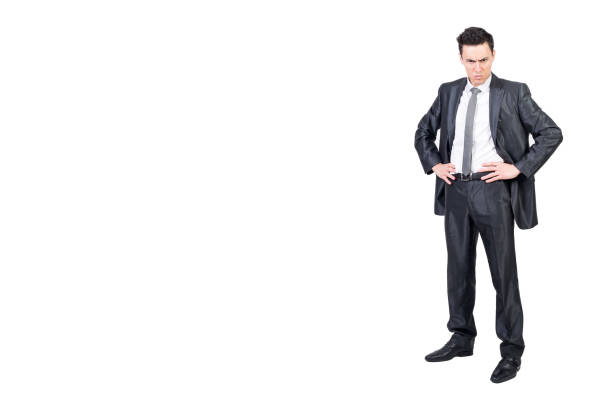 Full length of irritated male entrepreneur in formal suit standing with hands on waist and looking at camera with anger on white background