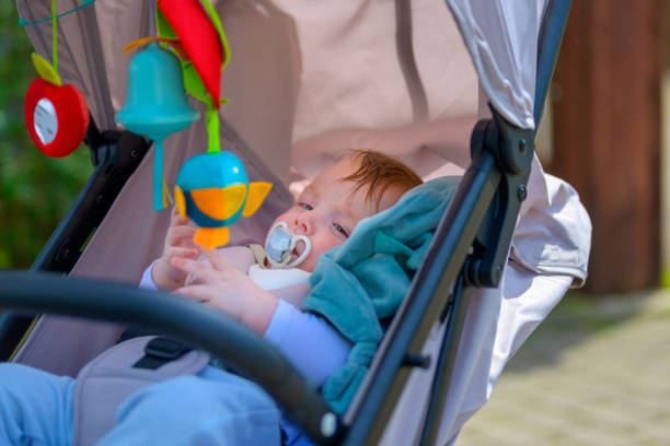 A baby with a pacifier relaxes in a stroller, engaging with colorful hanging toys on a sunny day.