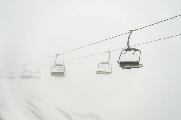 Two skiers are seated on a chairlift, heading through a snowy mountain winter scene. The image exudes feelings of adventure, leisure, and excitement in a serene and cold environment. The surrounding area is covered in snow, creating a perfect setting for winter sports and enjoying time in the mountains during the colder season.