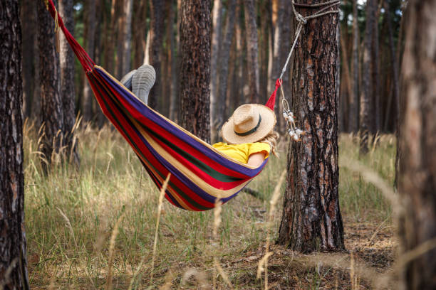 Sleeping woman with covered face by hat resting in hammock in forest. Napping and relaxing outdoors