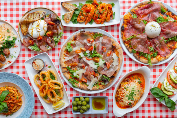 A tabletop view of an Italian restaurant dishes on a red and white checkered table cloth, dishes of a pizza with ham and broccoli, pizza with burrata and beef bresaola, fried calamari, burrata plate, goat’s cheese plate with avocados, green olives with olive oil, spaghetti Bolognese, shrimp, lasagna.