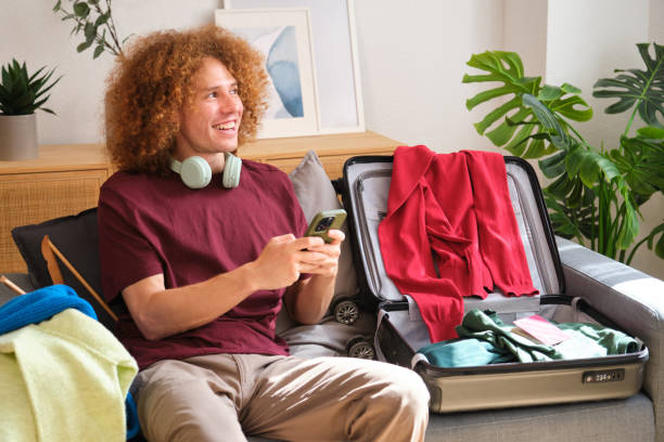 istock Happy young man using smartphone while packing suitcase for travel vacation 2160982987