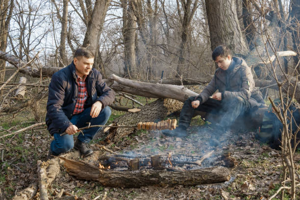 istock Two men make a campfire for cooking in the forest, father and son sitting on a log while hiking and outdoor activities, early spring landscape 2160991630
