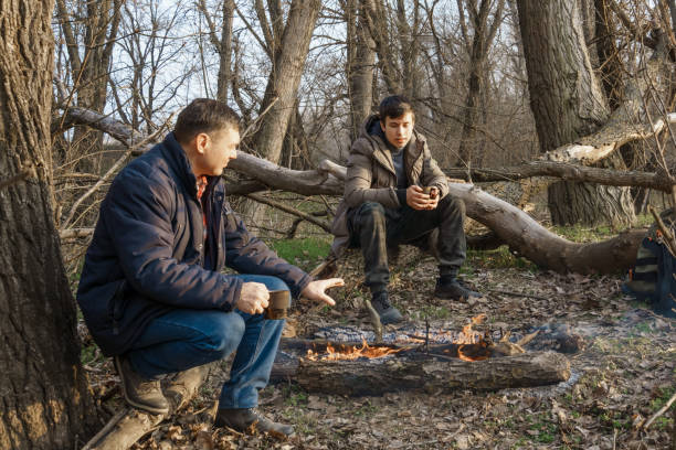 istock Two men make a campfire for cooking in the forest, father and son sitting on a log while hiking and outdoor activities, early spring landscape 2160991725