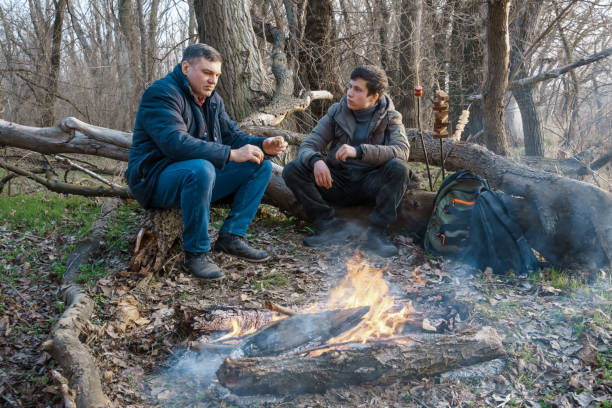 istock Two men make a campfire for cooking in the forest, father and son sitting on a log while hiking and outdoor activities, early spring landscape 2160991830