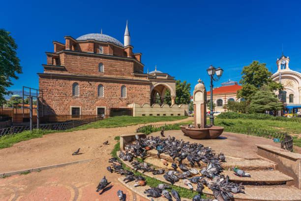 A flock of city pigeons by a water fountian in a town square, in the background the Banya Bashi Mosque and the Central Market of Sofia, Bulgaria