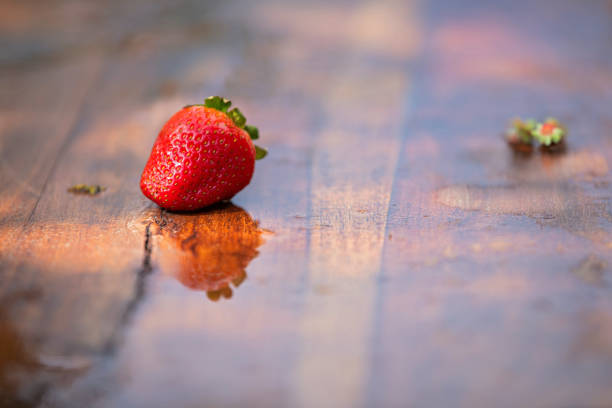 A red strawberry is sitting on a wooden table. The reflection of the strawberry can be seen in the water