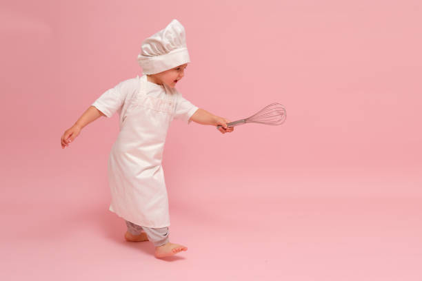 istock A child in a culinary outfit, including a chef's hat and apron, grips a whisk while posing in front of a pink backdrop. Kid aged 3 years (three year old boy) 2161033051