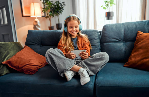 Concept of online activity. Relaxed blonde girl in headphones sitting on sofa with smartphone in hands Concept of online activity. Relaxed blonde girl in wireless headphones sitting on blue sofa with smartphone in hands. Interested kid spending time at home while playing video game on personal gadget. free girl download stock pictures, royalty-free photos & images