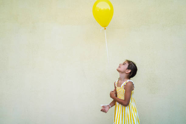A young girl in a yellow and white striped dress holds a yellow balloon while looking up, standing against a plain wall backdrop, capturing a joyful and playful moment.