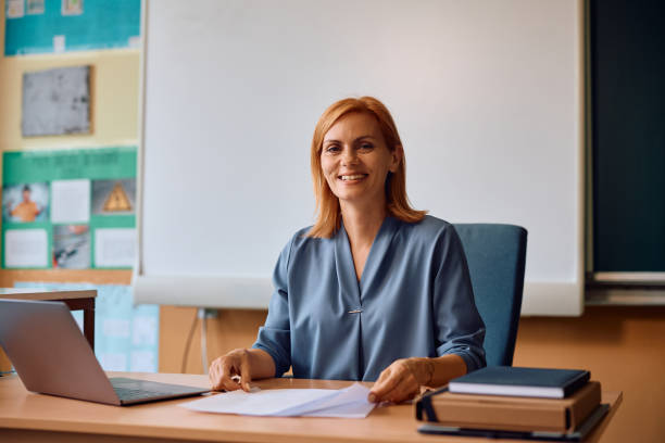 istock Happy teacher at her desk in the classroom looking at camera. 2161063593