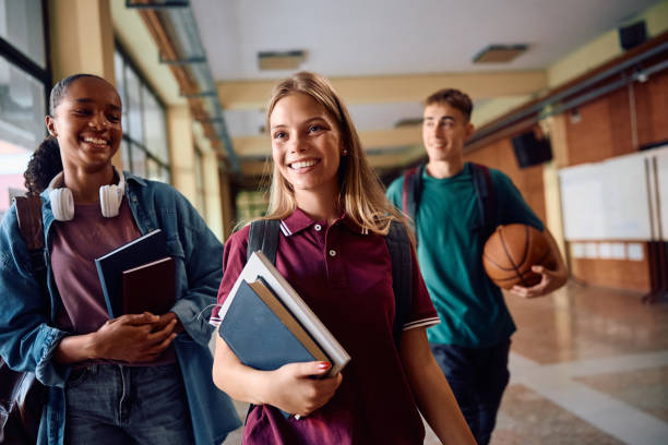Two students are walking outside university building, holding books and a soccer ball and talking to each other