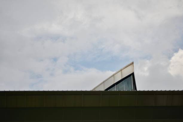 A photograph of the upper part of a modern building with a triangular glass structure extending above a flat roofline. The background features a partly cloudy sky, creating a contrast between the architectural elements and the natural setting.