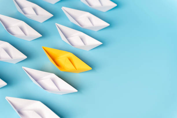 istock Yellow paper boat floating away from others on light background. 2161085446