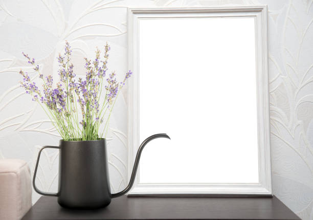 A black kettle with lavender flowers sits on a table next to a white picture frame. The background is a white wall with an embossed pattern.