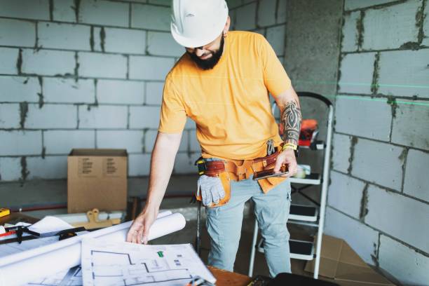 istock Mid adult construction worker in hard hat reading blueprints, using digital tablet on construction site 2161120886
