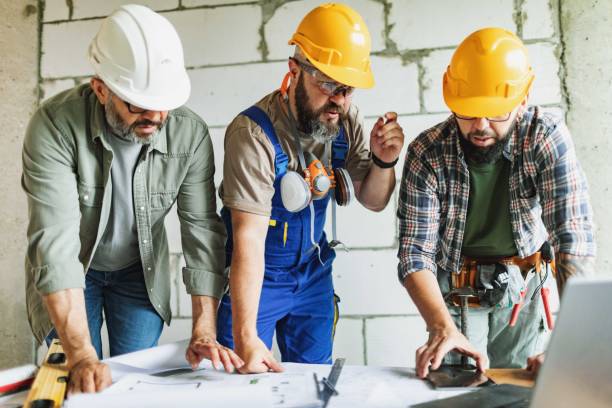 istock Three manual construction workers in hard hats discussing blueprints at construction site 2161121235
