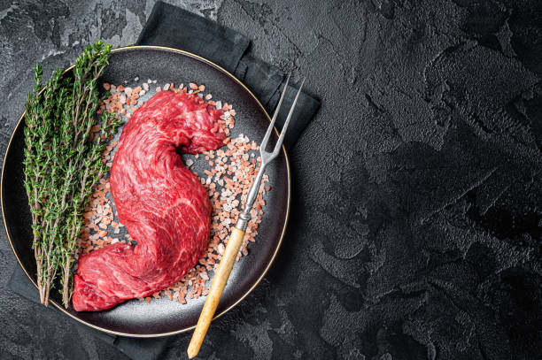 Raw Machete beef meat steak or hanging tender cut on plate with sea salt and thyme. Black background. Top view. Copy space