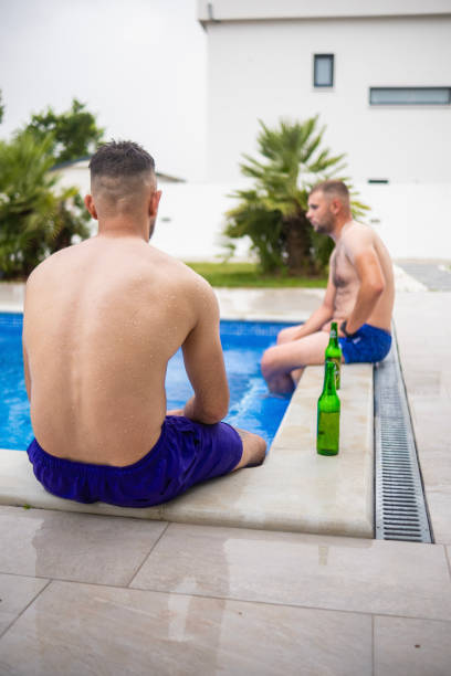 A man is sitting in a pool with a beer in his hand. The sky is darkening and the man is looking out over the water